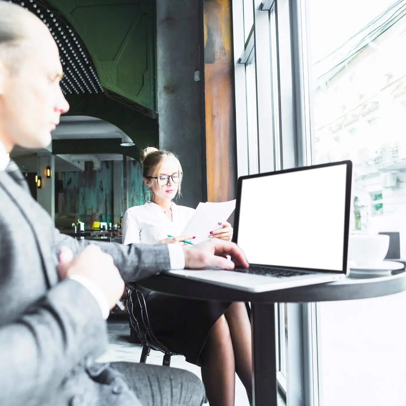 Geschäftsmann benutzt einen Laptop, während sein Kollege im Café Dokumente prüft.