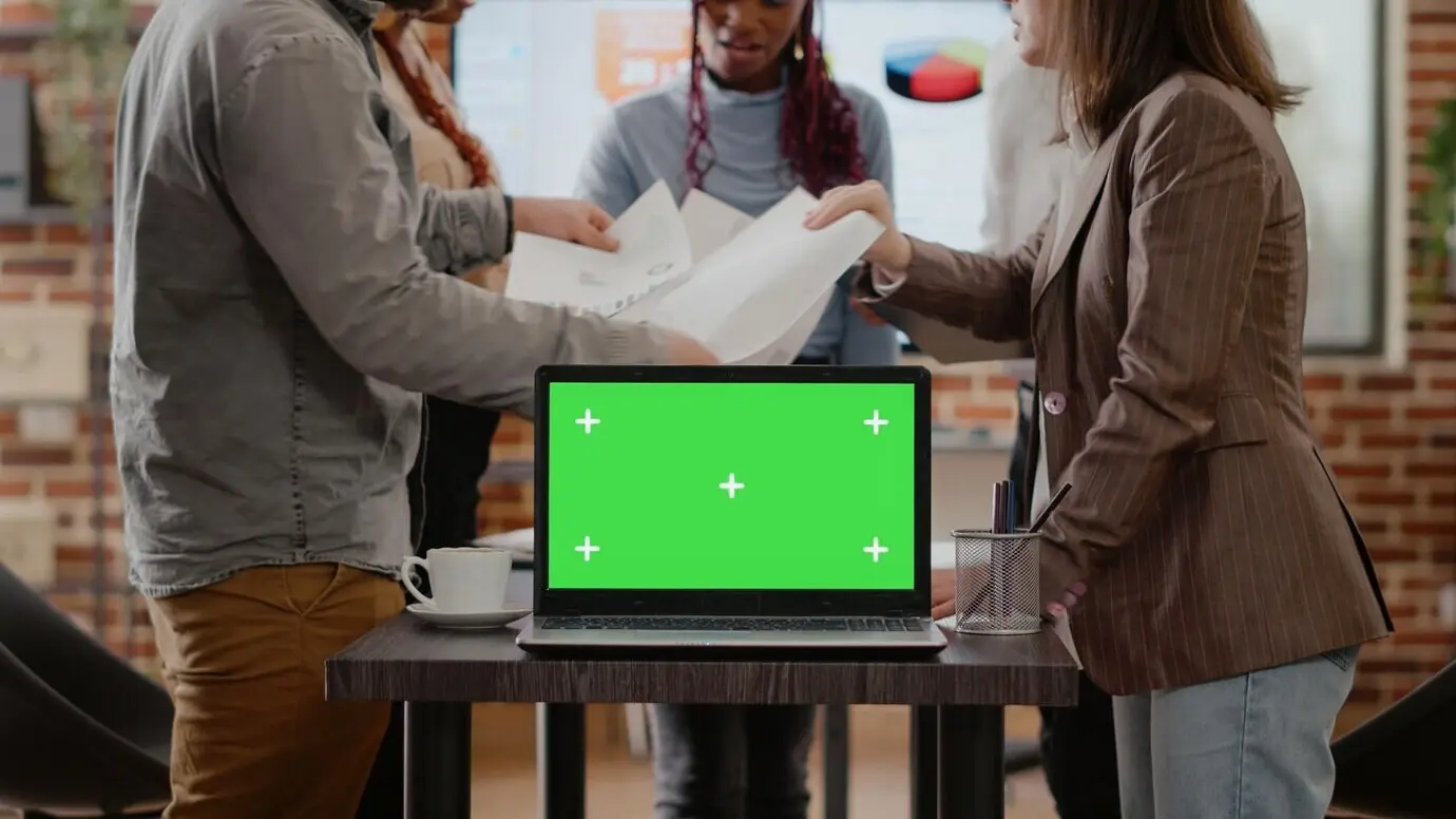 Nahaufnahme von Kollegen mit einem Laptop mit grünem Bildschirm auf dem Schreibtisch, die mit Dokumenten arbeiten; der Computer zeigt einen isolierten Chroma-Key-Copy-Space. Kollegen verwenden eine leere Mock-up-Vorlage und einen Hintergrund.